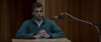 Movie still from “The Face of an Angel” (2014), directed by Michael Winterbottom – A man sitting in front of a microphone at a table; Medium shot, Over the shoulder angle