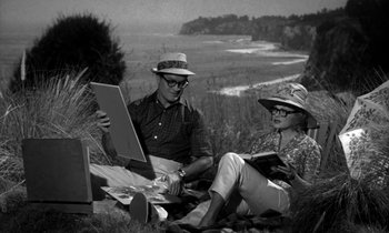 Movie still from “The Facts of Life” (1960), directed by Melvin Frank – A man and a woman sitting on the ground near a body of water; Wide shot, High angle