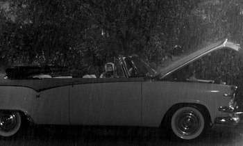 Movie still from “The Facts of Life” (1960), directed by Melvin Frank – An old car is parked on the side of the road in the pouring rain; Wide shot, High angle