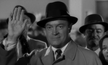 Movie still from “The Facts of Life” (1960), directed by Melvin Frank – A man wearing a hat and a trench coat waving; Close Up shot, Over the shoulder angle