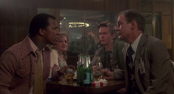 Movie still from “The Falcon and the Snowman” (1985), directed by John Schlesinger – A group of people sitting at a table with drinks on it; Medium shot, Over the shoulder angle