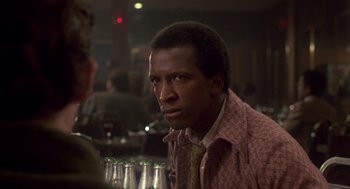 Movie still from “The Falcon and the Snowman” (1985), directed by John Schlesinger – A man sitting at a table in front of a group of people; Close Up shot, Over the shoulder angle