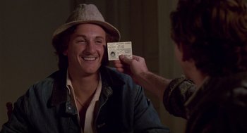 Movie still from “The Falcon and the Snowman” (1985), directed by John Schlesinger – A man holding up a photo of a man wearing a hat; Close Up shot, Over the shoulder angle
