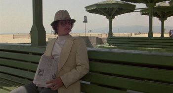 Movie still from “The Falcon and the Snowman” (1985), directed by John Schlesinger – A man wearing a hat and holding a newspaper on top of a park bench; Medium shot, Low angle