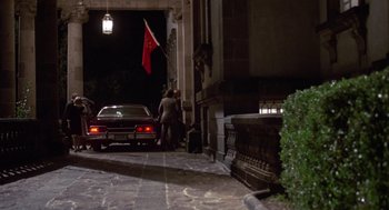 Movie still from “The Falcon and the Snowman” (1985), directed by John Schlesinger – Two people standing next to a car on the sidewalk; Wide shot, Over the shoulder angle