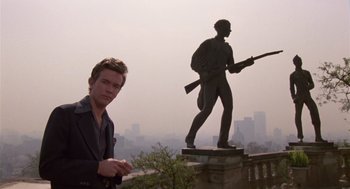 Movie still from “The Falcon and the Snowman” (1985), directed by John Schlesinger – A man standing next to a statue of a man holding a rifle; Medium shot, Low angle