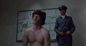 Movie still from “The Falcon and the Snowman” (1985), directed by John Schlesinger – A shirtless man standing in front of a police officer; Medium shot, Low angle