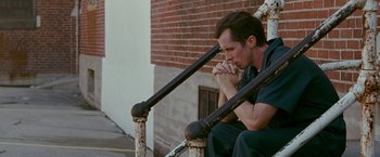 Movie still from “The Fighter” (2010), directed by David O. Russell – A man sitting on a railing with his hands clasped together; Medium shot, High angle