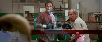 Movie still from “The Fighter” (2010), directed by David O. Russell – A man in a boxing ring holding a pair of boxing mitts; Medium shot, Over the shoulder angle