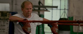 Movie still from “The Fighter” (2010), directed by David O. Russell – An older man wearing a white t - shirt is holding a pair of boxing mitts; Close Up shot, Low angle