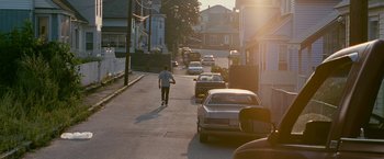Movie still from “The Fighter” (2010), directed by David O. Russell – A man riding a skateboard down the side of a street; Wide shot, Over the shoulder angle