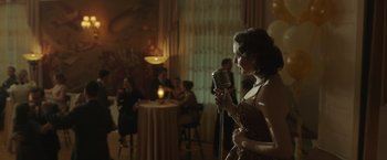 Movie still from “The Finest Hours” (2016), directed by Craig Gillespie – A woman holding a microphone in front of a crowd; Medium shot, Over the shoulder angle