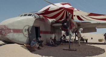 Movie still from “The Flight of the Phoenix” (1965), directed by Robert Aldrich – A group of people sitting under an awning next to an airplane; Extreme Wide shot, Low angle