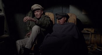 Movie still from “The Flight of the Phoenix” (1965), directed by Robert Aldrich – Two men sitting on a chair in a dark room; Medium shot, High angle