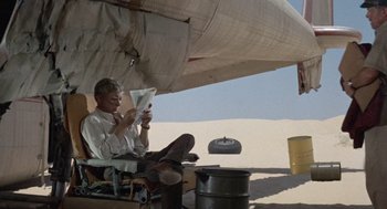 Movie still from “The Flight of the Phoenix” (1965), directed by Robert Aldrich – A man sitting on the ground reading a book in the desert; Wide shot, High angle