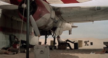 Movie still from “The Flight of the Phoenix” (1965), directed by Robert Aldrich – A man sitting under an airplane on the ground; Wide shot, Low angle