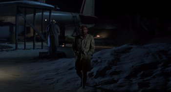 Movie still from “The Flight of the Phoenix” (1965), directed by Robert Aldrich – A man standing in the snow next to an airplane at night; Wide shot, High angle