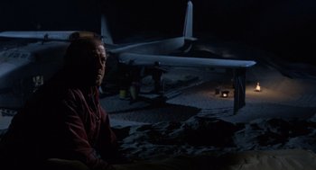 Movie still from “The Flight of the Phoenix” (1965), directed by Robert Aldrich – A man sitting in front of an airplane at night; Wide shot, Low angle