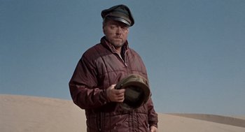 Movie still from “The Flight of the Phoenix” (1965), directed by Robert Aldrich – A man wearing a hat standing in the sand; Medium shot, Over the shoulder angle