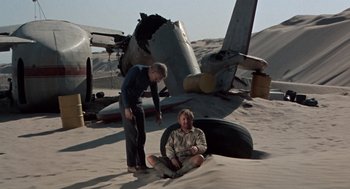Movie still from “The Flight of the Phoenix” (1965), directed by Robert Aldrich – Two men are sitting in the sand next to an airplane wreck; Wide shot, High angle