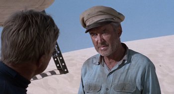 Movie still from “The Flight of the Phoenix” (1965), directed by Robert Aldrich – An older man wearing a hat standing in the sand; Close Up shot, Over the shoulder angle