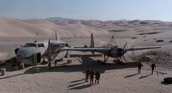 Movie still from “The Flight of the Phoenix” (1965), directed by Robert Aldrich – An airplane is parked in the sand with people standing around it; Extreme Wide shot, Low angle