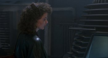 Movie still from “The Fly” (1986), directed by David Cronenberg – A woman with curly hair is standing in a dark room; Medium shot, Over the shoulder angle