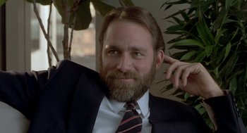 Movie still from “The Fly” (1986), directed by David Cronenberg – A man in a suit and tie sitting in front of a plant; Close Up shot, Over the shoulder angle