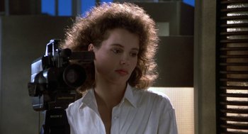 Movie still from “The Fly” (1986), directed by David Cronenberg – A woman in a white dress shirt is standing in front of a video camera; Close Up shot, Low angle