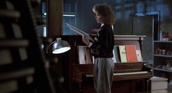 Movie still from “The Fly” (1986), directed by David Cronenberg – A woman standing in front of a piano holding a sheet; Medium shot, Over the shoulder angle