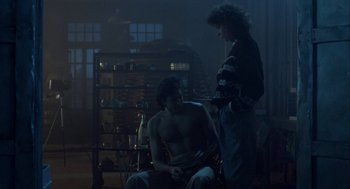 Movie still from “The Fly” (1986), directed by David Cronenberg – A man sitting in a chair next to a woman; Medium shot, Over the shoulder angle
