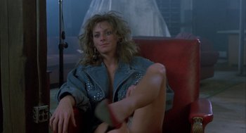 Movie still from “The Fly” (1986), directed by David Cronenberg – A woman sitting on a red couch in front of a microphone; Medium shot, Low angle