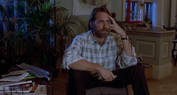 Movie still from “The Fly” (1986), directed by David Cronenberg – A man sitting in a chair in a living room; Medium shot, Low angle