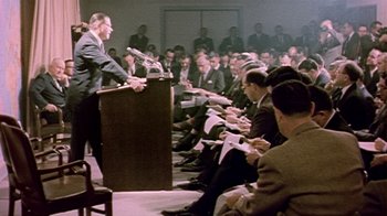 Movie still from “The Fog of War” (2003), directed by Errol Morris – A man standing at a podium in front of a group of people; Medium shot, Over the shoulder angle