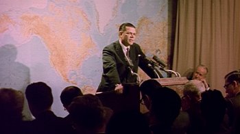 Movie still from “The Fog of War” (2003), directed by Errol Morris – A man standing at a podium speaking to a crowd; Medium shot, Over the shoulder angle