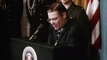Movie still from “The Fog of War” (2003), directed by Errol Morris – A man in a suit and glasses is speaking at a podium; Close Up shot, Low angle