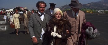 Movie still from “The Fortune” (1975), directed by Mike Nichols – A woman in a fur coat and a hat walking with a man in a suit and tie; Medium shot, Over the shoulder angle