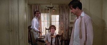 Movie still from “The Fortune” (1975), directed by Mike Nichols – A woman sitting at a dining room table with two men standing behind her; Medium shot, Over the shoulder angle