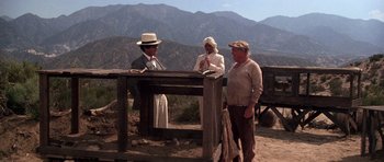 Movie still from “The Fortune” (1975), directed by Mike Nichols – Two men and a woman standing in front of a wooden table; Wide shot, High angle