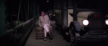 Movie still from “The Fortune” (1975), directed by Mike Nichols – A man sitting on top of a piece of luggage on the ground; Wide shot, High angle