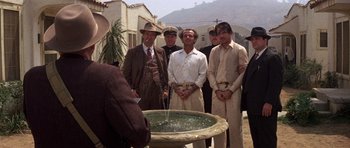 Movie still from “The Fortune” (1975), directed by Mike Nichols – A group of men standing next to each other in front of a fountain; Wide shot, Over the shoulder angle