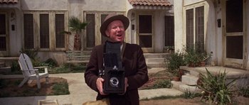 Movie still from “The Fortune” (1975), directed by Mike Nichols – A man holding an old fashioned camera in front of a house; Close Up shot, Low angle
