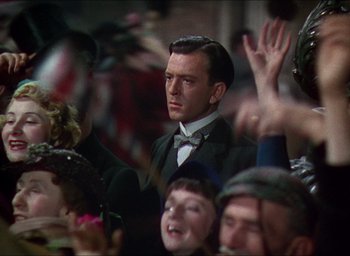Movie still from “The Four Feathers” (1939), directed by Zoltan Korda – A man wearing a suit and bow tie in front of a group of people; Close Up shot, Over the shoulder angle