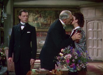 Movie still from “The Four Feathers” (1939), directed by Zoltan Korda – Two men and a woman kissing in front of a table with flowers; Medium shot, Over the shoulder angle