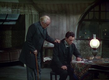 Movie still from “The Four Feathers” (1939), directed by Zoltan Korda – A man sitting at a table with another man standing next to him; Medium shot, High angle