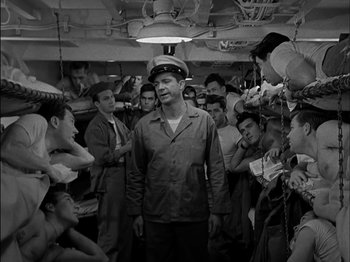 Movie still from “The Frogmen” (1951), directed by Lloyd Bacon – A group of people gathered around a man wearing a hat; Medium shot, High angle