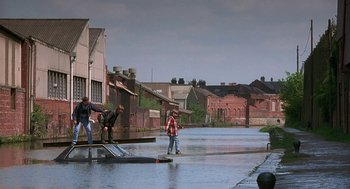 Movie still from “The Full Monty” (1997), directed by Peter Cattaneo – A group of people standing on top of a flooded street; Extreme Wide shot, High angle