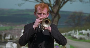 Movie still from “The Full Monty” (1997), directed by Peter Cattaneo – A man in a black suit and red hair blowing into a trumpet; Medium shot, Low angle