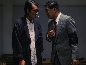 Two men in suits smoking a cigarette and talking to each other