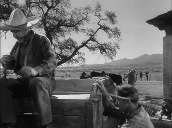 Movie still from “The Furies” (1950), directed by Anthony Mann – A black and white photo of a man sitting on top of a wooden bench; Wide shot, Low angle